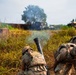 Military personnel in camouflage uniforms and tactical gear are operating a mortar system in an open field environment. Smoke is visible rising from the mortar tube, indicating a recent or ongoing firing sequence. Military vehicles are visible in the background amid tall grass and trees.