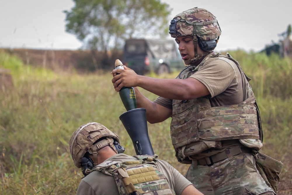 Two military personnel in full combat gear are conducting a mortar operation in a field environment. The standing soldier is preparing to drop a mortar round into a mortar tube held by the second soldier on the ground. The setting appears to be a training exercise or field operation in a tropical or subtropical environment.