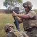 Two military personnel in full combat gear are conducting a mortar operation in a field environment. The standing soldier is preparing to drop a mortar round into a mortar tube held by the second soldier on the ground. The setting appears to be a training exercise or field operation in a tropical or subtropical environment.