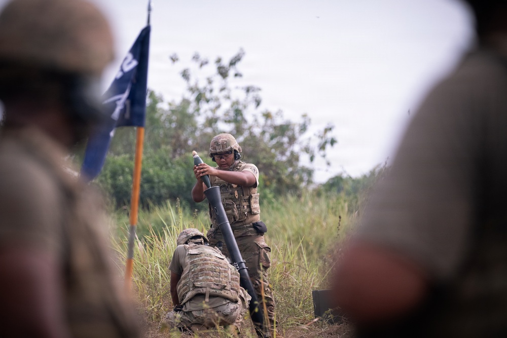 A military training exercise is depicted with soldiers in multicam/OCP camouflage uniforms operating a mortar system in a field environment. A standing soldier is preparing to drop a mortar round into the tube while a second soldier kneels beside the emplaced mortar. A flag on a pole, appearing to have blue and orange/yellow colors, is visible in the background.