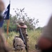A military training exercise is depicted with soldiers in multicam/OCP camouflage uniforms operating a mortar system in a field environment. A standing soldier is preparing to drop a mortar round into the tube while a second soldier kneels beside the emplaced mortar. A flag on a pole, appearing to have blue and orange/yellow colors, is visible in the background.