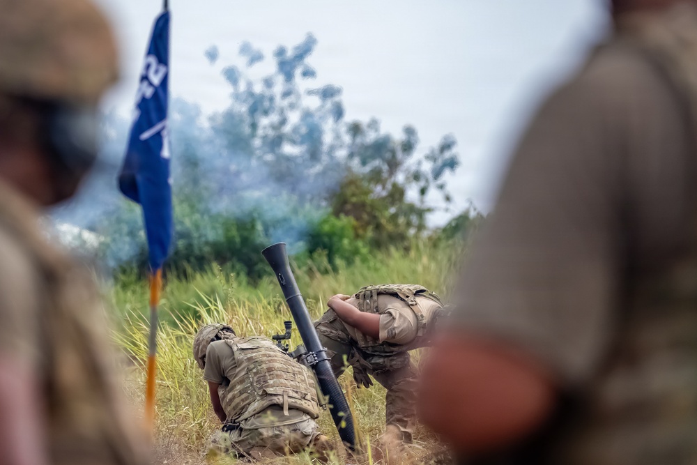 The image shows military personnel in camouflage uniforms operating what appears to be a mortar system in a field setting. Smoke is visible in the background, suggesting a recent or ongoing firing exercise. A blue military unit guidon flag is visible to the left, indicating this is likely a formal training exercise.
