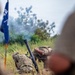 The image shows military personnel in camouflage uniforms operating what appears to be a mortar system in a field setting. Smoke is visible in the background, suggesting a recent or ongoing firing exercise. A blue military unit guidon flag is visible to the left, indicating this is likely a formal training exercise.