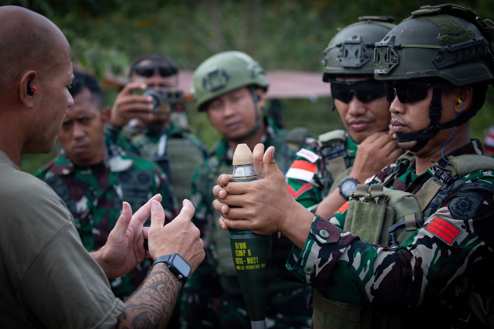 A military training or demonstration session is taking place outdoors, where a civilian-dressed instructor appears to be briefing a group of soldiers in camouflage uniforms. One soldier is holding a munition canister labeled '81MM COMP B ERTG-M827', while another documents the session with a camera. The soldiers appear to be from the Indonesian Armed Forces (TNI), identifiable by their camouflage pattern and Indonesian flag patches.