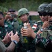 A military training or demonstration session is taking place outdoors, where a civilian-dressed instructor appears to be briefing a group of soldiers in camouflage uniforms. One soldier is holding a munition canister labeled '81MM COMP B ERTG-M827', while another documents the session with a camera. The soldiers appear to be from the Indonesian Armed Forces (TNI), identifiable by their camouflage pattern and Indonesian flag patches.