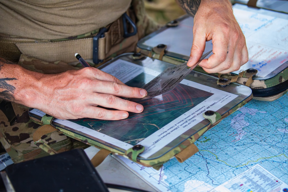 A person in military camouflage uniform is working at a field operations station, examining documents on a tactical clipboard or map board. The individual appears to be conducting mission planning or navigation work, using what appears to be a printed aerial or topographic image overlaid with a transparent acetate sheet. Multiple maps and documents are visible on the work surface.