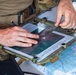 A person in military camouflage uniform is working at a field operations station, examining documents on a tactical clipboard or map board. The individual appears to be conducting mission planning or navigation work, using what appears to be a printed aerial or topographic image overlaid with a transparent acetate sheet. Multiple maps and documents are visible on the work surface.
