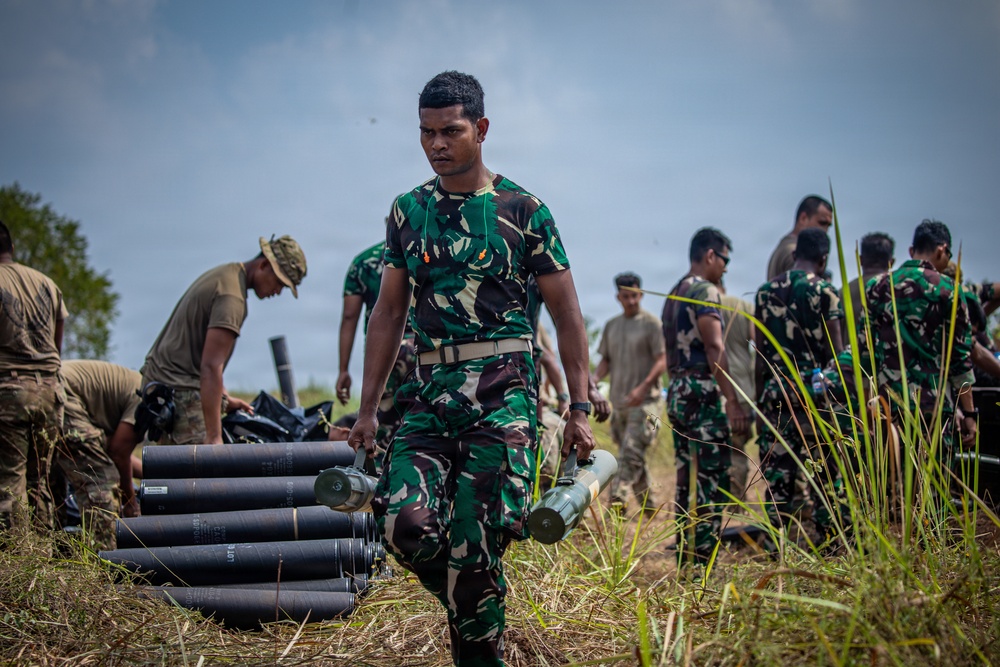 The image shows a group of military personnel, predominantly wearing green camouflage uniforms consistent with Indonesian Army (TNI-AD) pattern, conducting what appears to be an artillery or ordnance handling exercise in an outdoor field setting. A central soldier carries two projectile casings while others handle similar munitions in the background. At least one soldier in a different uniform (tan/multicam) is also visible, suggesting a joint or multinational training exercise.