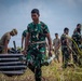 The image shows a group of military personnel, predominantly wearing green camouflage uniforms consistent with Indonesian Army (TNI-AD) pattern, conducting what appears to be an artillery or ordnance handling exercise in an outdoor field setting. A central soldier carries two projectile casings while others handle similar munitions in the background. At least one soldier in a different uniform (tan/multicam) is also visible, suggesting a joint or multinational training exercise.