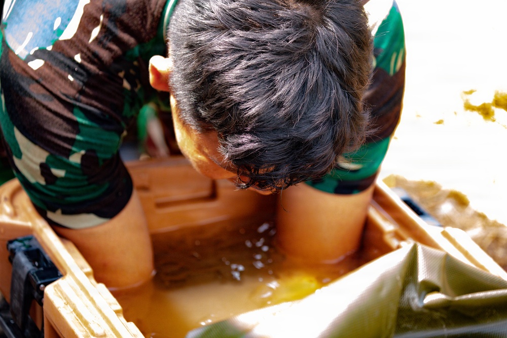 A person wearing a camouflage uniform is leaning forward with their face submerged or very close to a brown liquid-filled container. The container appears to be a plastic tub or cooler filled with murky brown/amber-colored water or liquid. This appears to be a physical challenge or training exercise, possibly a military or obstacle course activity.