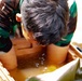 A person wearing a camouflage uniform is leaning forward with their face submerged or very close to a brown liquid-filled container. The container appears to be a plastic tub or cooler filled with murky brown/amber-colored water or liquid. This appears to be a physical challenge or training exercise, possibly a military or obstacle course activity.