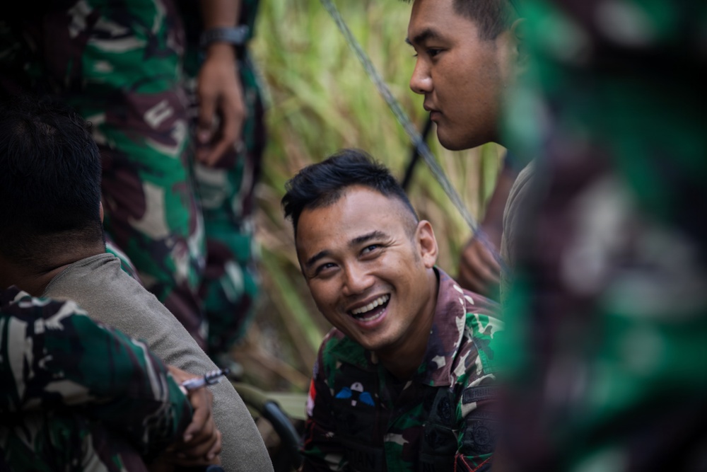 A group of military personnel wearing green camouflage uniforms are gathered together in an outdoor setting with vegetation in the background. One soldier in the center is laughing broadly, suggesting a moment of camaraderie or humor during a break. The image captures an informal, candid moment among what appear to be Southeast Asian military personnel.