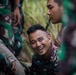 A group of military personnel wearing green camouflage uniforms are gathered together in an outdoor setting with vegetation in the background. One soldier in the center is laughing broadly, suggesting a moment of camaraderie or humor during a break. The image captures an informal, candid moment among what appear to be Southeast Asian military personnel.