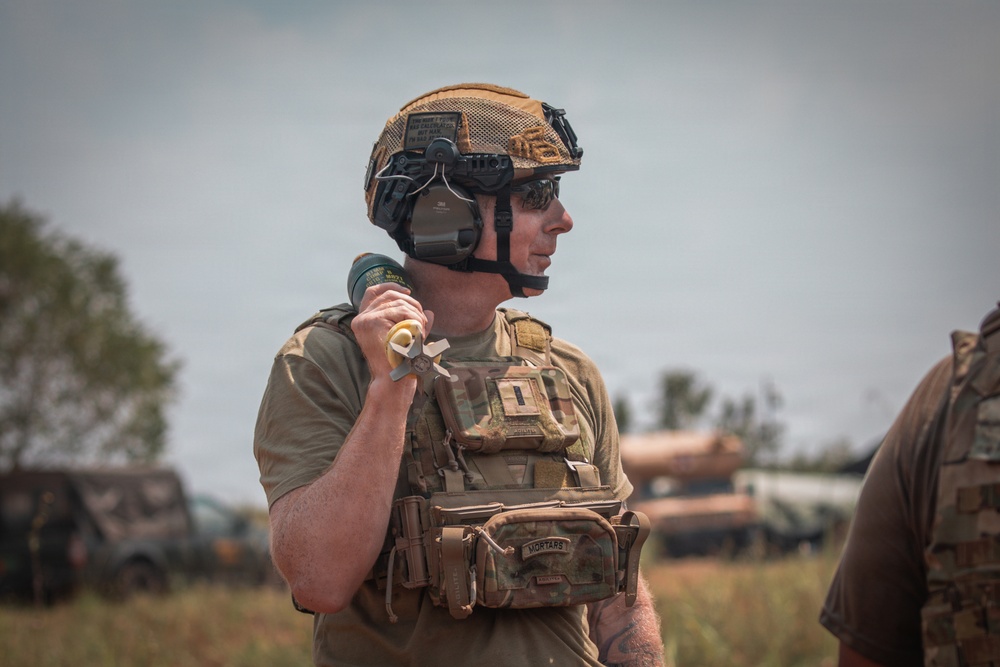A male soldier in full tactical gear is photographed in an outdoor field environment. He is wearing a tan/coyote brown ballistic helmet with communications equipment and a multicam plate carrier vest with a labeled 'MORTARS' pouch. He appears to be holding a mortar round or similar munition in his right hand.