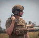 A male soldier in full tactical gear is photographed in an outdoor field environment. He is wearing a tan/coyote brown ballistic helmet with communications equipment and a multicam plate carrier vest with a labeled 'MORTARS' pouch. He appears to be holding a mortar round or similar munition in his right hand.