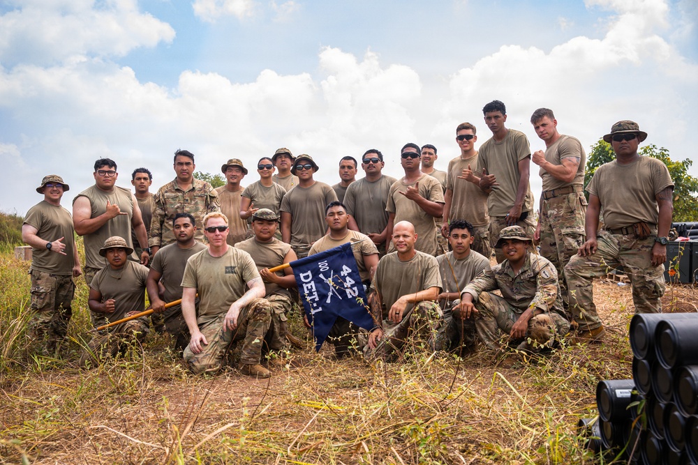 A group of approximately 20-25 military personnel in camouflage uniforms pose together for a group photo outdoors in a field setting. They are holding a blue unit guidon/pennant that reads '100/442 DET-1', referencing the historic 100th Battalion/442nd Infantry Regiment. The photo appears to be taken during a training exercise or field operation under partly cloudy skies.