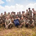 A group of approximately 20-25 military personnel in camouflage uniforms pose together for a group photo outdoors in a field setting. They are holding a blue unit guidon/pennant that reads '100/442 DET-1', referencing the historic 100th Battalion/442nd Infantry Regiment. The photo appears to be taken during a training exercise or field operation under partly cloudy skies.