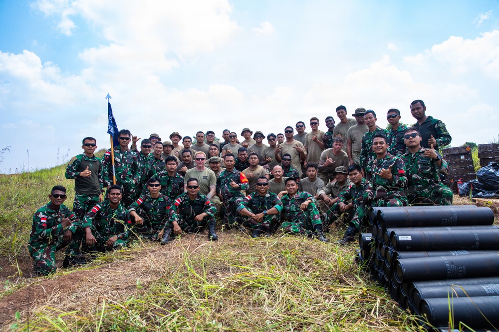 A large group of military personnel from two distinct forces pose together for a group photograph in an outdoor field setting. The group includes soldiers wearing green camouflage uniforms (consistent with Indonesian TNI/Army pattern) and others in tan/khaki uniforms. Several cylindrical black tubes are stacked in the foreground, and a blue military guidon/flag is visible on the left side.