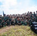 A large group of military personnel from two distinct forces pose together for a group photograph in an outdoor field setting. The group includes soldiers wearing green camouflage uniforms (consistent with Indonesian TNI/Army pattern) and others in tan/khaki uniforms. Several cylindrical black tubes are stacked in the foreground, and a blue military guidon/flag is visible on the left side.