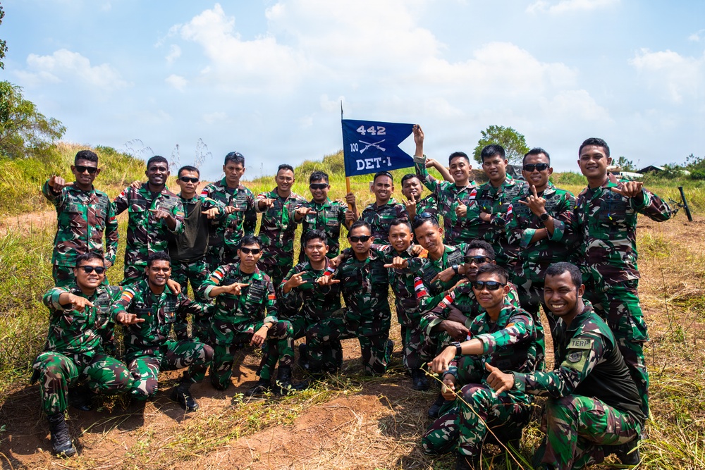 A group of approximately 20 military personnel in camouflage uniforms pose together outdoors for a group photograph. They are holding a blue pennant flag marked '442 / 100 / DET-1' with crossed rifles insignia. The soldiers appear to be in a celebratory mood, making hand gestures and smiling.
