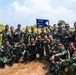 A group of approximately 20 military personnel in camouflage uniforms pose together outdoors for a group photograph. They are holding a blue pennant flag marked '442 / 100 / DET-1' with crossed rifles insignia. The soldiers appear to be in a celebratory mood, making hand gestures and smiling.