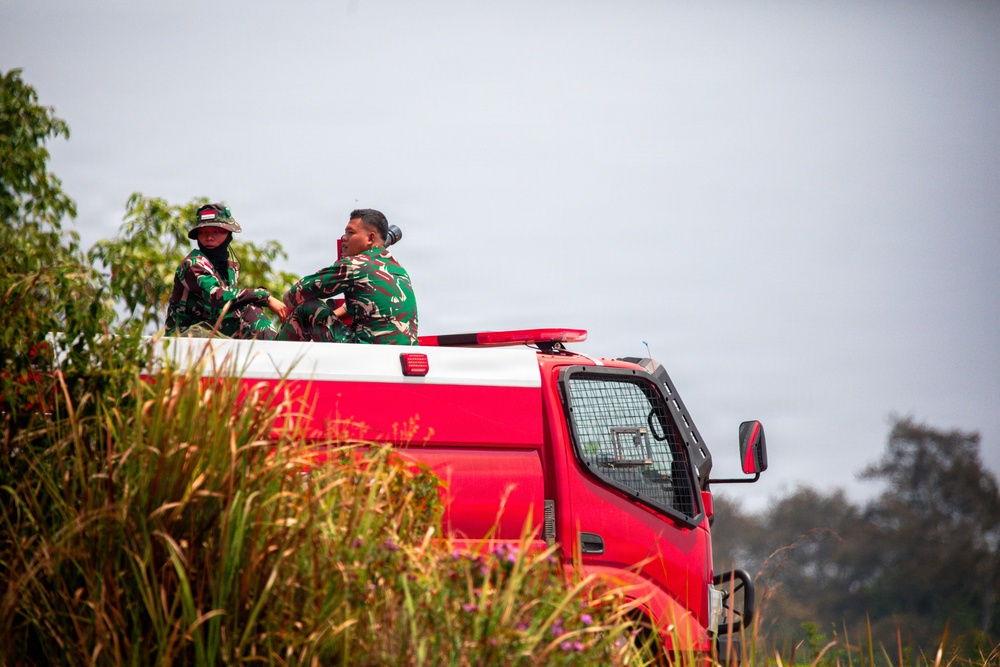 Two individuals wearing Indonesian military (TNI) camouflage uniforms are riding in the back of a red fire truck or emergency response vehicle. They appear to be on patrol or responding to an incident, surrounded by vegetation suggesting a rural or semi-rural environment. The Indonesian flag patch is visible on at least one uniform.