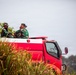 Two individuals wearing Indonesian military (TNI) camouflage uniforms are riding in the back of a red fire truck or emergency response vehicle. They appear to be on patrol or responding to an incident, surrounded by vegetation suggesting a rural or semi-rural environment. The Indonesian flag patch is visible on at least one uniform.