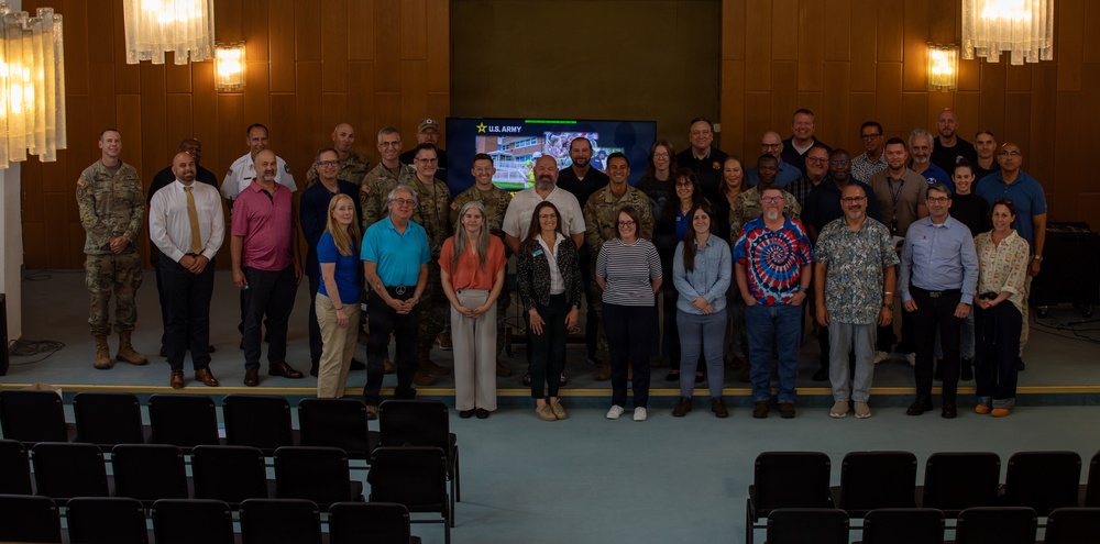 A large group of approximately 35-40 individuals poses for a formal group photograph in what appears to be an auditorium or conference hall. The group consists of a mix of military personnel in camouflage uniforms and civilians in business casual attire. A U.S. Army branded display screen is visible in the background, suggesting this is an official military or government-related event.