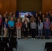 A large group of approximately 35-40 individuals poses for a formal group photograph in what appears to be an auditorium or conference hall. The group consists of a mix of military personnel in camouflage uniforms and civilians in business casual attire. A U.S. Army branded display screen is visible in the background, suggesting this is an official military or government-related event.