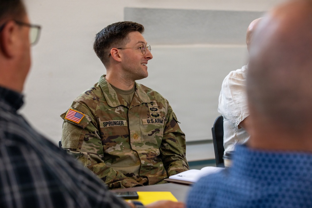 A U.S. Army soldier named Sprunger is seated at a table in what appears to be a briefing or meeting room, smiling and engaged in conversation. He is wearing the Operational Camouflage Pattern (OCP) uniform with a U.S. flag patch, name tape, and U.S. Army tape visible. Two other individuals, out of focus in the foreground, are also present at the meeting.