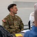 A U.S. Army soldier named Sprunger is seated at a table in what appears to be a briefing or meeting room, smiling and engaged in conversation. He is wearing the Operational Camouflage Pattern (OCP) uniform with a U.S. flag patch, name tape, and U.S. Army tape visible. Two other individuals, out of focus in the foreground, are also present at the meeting.