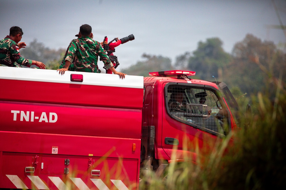 A red military fire truck marked 'TNI-AD' (Tentara Nasional Indonesia - Angkatan Darat, Indonesian Army) is shown with personnel in green camouflage uniforms riding on top. One soldier is operating or aiming what appears to be a water cannon or firefighting nozzle mounted on the vehicle. The scene appears to be outdoors with vegetation visible in the background.