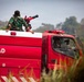 A red military fire truck marked 'TNI-AD' (Tentara Nasional Indonesia - Angkatan Darat, Indonesian Army) is shown with personnel in green camouflage uniforms riding on top. One soldier is operating or aiming what appears to be a water cannon or firefighting nozzle mounted on the vehicle. The scene appears to be outdoors with vegetation visible in the background.