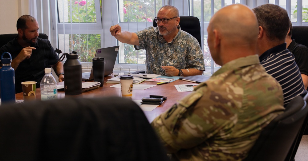 A meeting is taking place around a conference table with approximately four to five individuals, one of whom is wearing military camouflage uniform. A man in a floral/Hawaiian shirt appears to be leading the discussion, gesturing with his hand toward another participant. The setting appears to be an office with windows overlooking greenery.