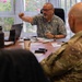 A meeting is taking place around a conference table with approximately four to five individuals, one of whom is wearing military camouflage uniform. A man in a floral/Hawaiian shirt appears to be leading the discussion, gesturing with his hand toward another participant. The setting appears to be an office with windows overlooking greenery.