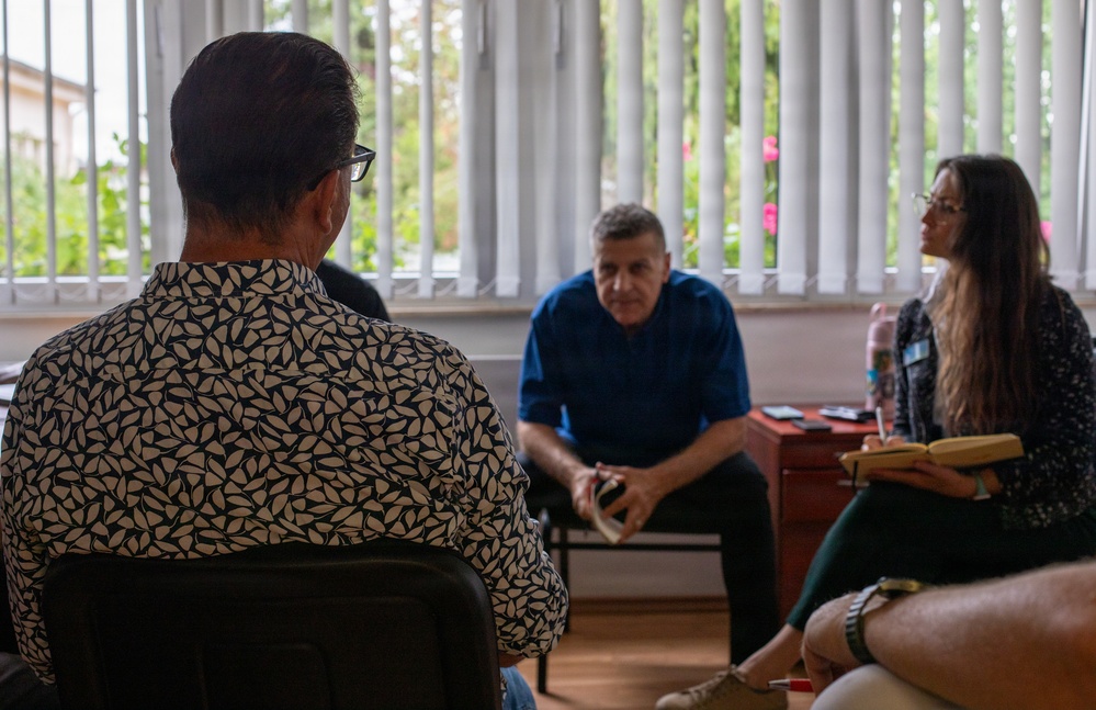 A small group of people are seated in a room engaged in what appears to be a meeting or interview session. The scene shows three clearly visible individuals arranged in a semi-circle, with one person photographed from behind in the foreground. The setting appears to be an indoor office or meeting room with vertical blinds and greenery visible through the window.