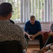 A small group of people are seated in a room engaged in what appears to be a meeting or interview session. The scene shows three clearly visible individuals arranged in a semi-circle, with one person photographed from behind in the foreground. The setting appears to be an indoor office or meeting room with vertical blinds and greenery visible through the window.