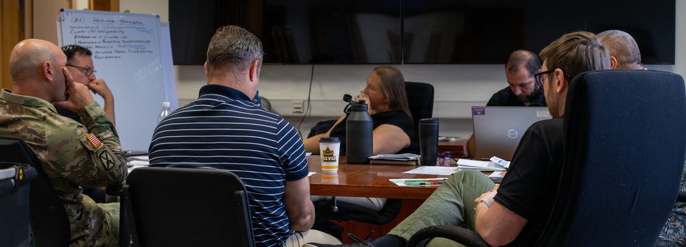 A group of approximately six individuals are seated around a conference table in what appears to be a professional meeting or working session. At least one participant is wearing U.S. Army military camouflage uniform, suggesting a military or government-related meeting. A whiteboard with handwritten notes is visible in the background alongside a large display screen.