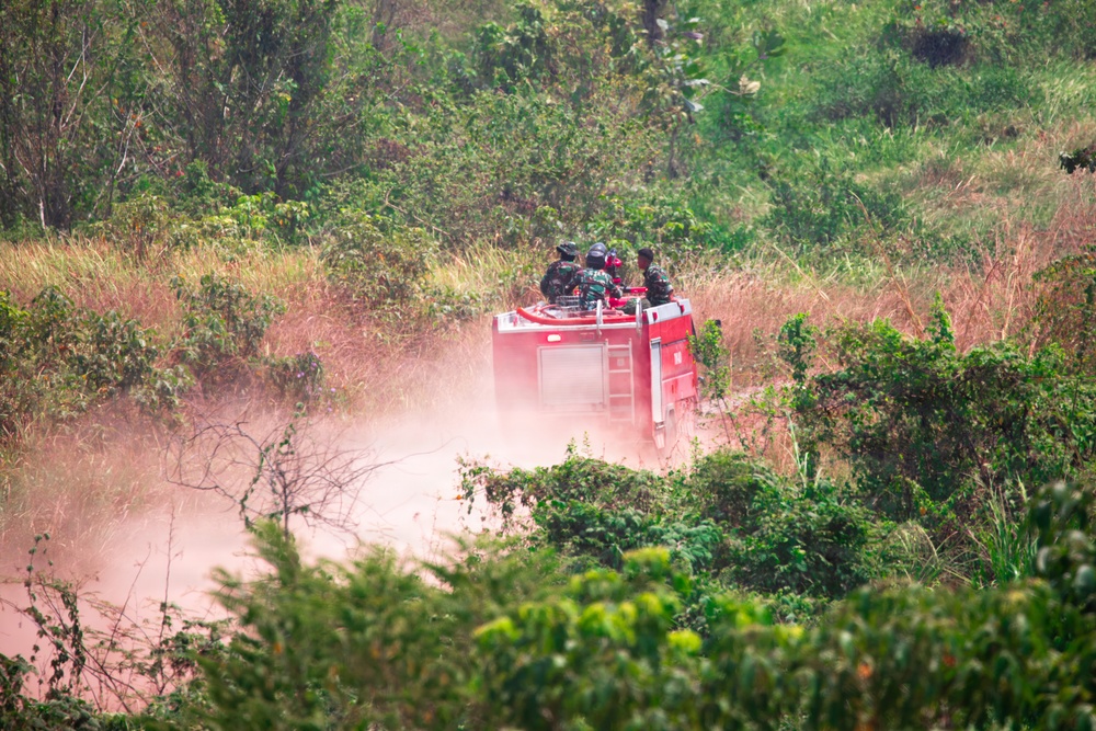 A red fire truck or emergency vehicle is traveling at speed along a dusty dirt road through dense tropical vegetation. Several individuals wearing what appear to be military or tactical uniforms are visible riding on top of the vehicle. The scene is set in a lush, green bush or jungle environment with reddish-brown dry grass mixed in.