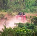 A red fire truck or emergency vehicle is traveling at speed along a dusty dirt road through dense tropical vegetation. Several individuals wearing what appear to be military or tactical uniforms are visible riding on top of the vehicle. The scene is set in a lush, green bush or jungle environment with reddish-brown dry grass mixed in.