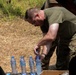 A man in military-style clothing (olive green t-shirt and camouflage pants) is leaning over a cardboard box containing multiple water bottles branded 'Club'. He appears to be opening or handling one of the water bottles in an outdoor field setting with dry grass and green vegetation in the background. Other individuals are partially visible at the edges of the frame.