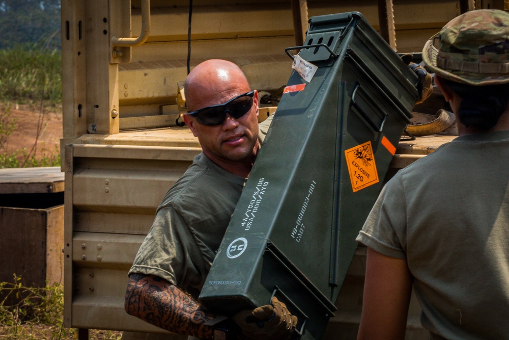 Two military personnel in olive drab t-shirts are unloading or transferring a large green ammunition crate from a tan military vehicle. The crate bears an orange explosive hazard label marked 'EXPLOSIVE 1.2G' along with military stenciling and serial numbers. The setting appears to be an outdoor training or operational area with red dirt terrain visible in the background.