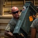 Two military personnel in olive drab t-shirts are unloading or transferring a large green ammunition crate from a tan military vehicle. The crate bears an orange explosive hazard label marked 'EXPLOSIVE 1.2G' along with military stenciling and serial numbers. The setting appears to be an outdoor training or operational area with red dirt terrain visible in the background.