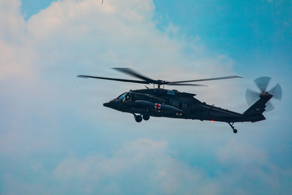 A dark military helicopter, identifiable as a UH-60 Black Hawk variant configured for medical evacuation (MEDEVAC), is captured in flight against a partly cloudy blue sky. The helicopter displays a prominent red cross symbol on its fuselage, indicating its medical/rescue role. The rotor blades show motion blur, confirming active flight.