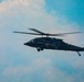 A dark military helicopter, identifiable as a UH-60 Black Hawk variant configured for medical evacuation (MEDEVAC), is captured in flight against a partly cloudy blue sky. The helicopter displays a prominent red cross symbol on its fuselage, indicating its medical/rescue role. The rotor blades show motion blur, confirming active flight.