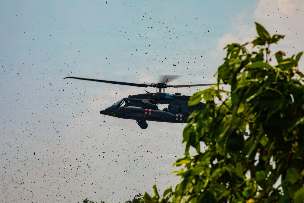 A military medical helicopter, identifiable by red cross markings on its fuselage, is captured in flight at low altitude near tree canopy. The rotor wash has disturbed significant debris, leaves, and particulate matter visible throughout the air around the aircraft. The helicopter appears to be a UH-60 Black Hawk variant configured for medevac/medical evacuation operations.