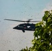 A military medical helicopter, identifiable by red cross markings on its fuselage, is captured in flight at low altitude near tree canopy. The rotor wash has disturbed significant debris, leaves, and particulate matter visible throughout the air around the aircraft. The helicopter appears to be a UH-60 Black Hawk variant configured for medevac/medical evacuation operations.