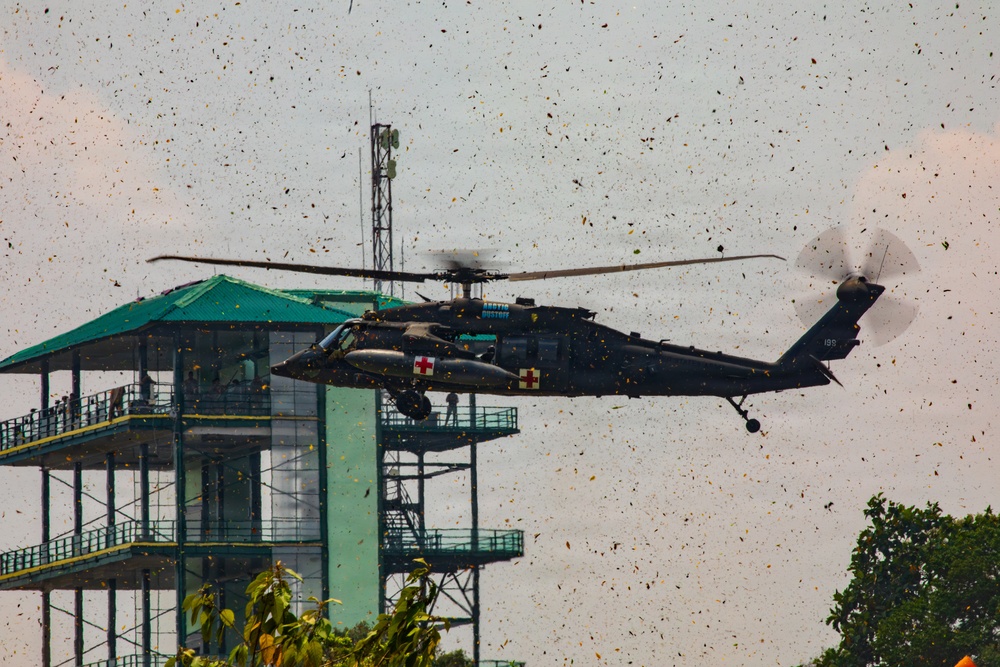 A military medical evacuation (MEDEVAC) helicopter, identifiable by the red cross markings on its fuselage, is hovering near a multi-story observation or control tower with a green roof. The helicopter's rotor wash has kicked up a large amount of debris, leaves, and particles filling the air around it. The text 'DUSTOFF' is partially visible on the aircraft, indicating it is a dedicated MEDEVAC unit.