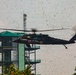 A military medical evacuation (MEDEVAC) helicopter, identifiable by the red cross markings on its fuselage, is hovering near a multi-story observation or control tower with a green roof. The helicopter's rotor wash has kicked up a large amount of debris, leaves, and particles filling the air around it. The text 'DUSTOFF' is partially visible on the aircraft, indicating it is a dedicated MEDEVAC unit.