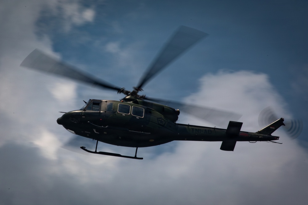 A military helicopter is captured in flight against a partly cloudy sky, photographed from a low angle looking upward. The aircraft appears to be a dark olive/green colored utility helicopter, likely a Bell 412 or similar variant, with rotor blades in motion creating visible motion blur. The image has a dramatic, low-angle perspective emphasizing the helicopter's silhouette against the overcast sky.